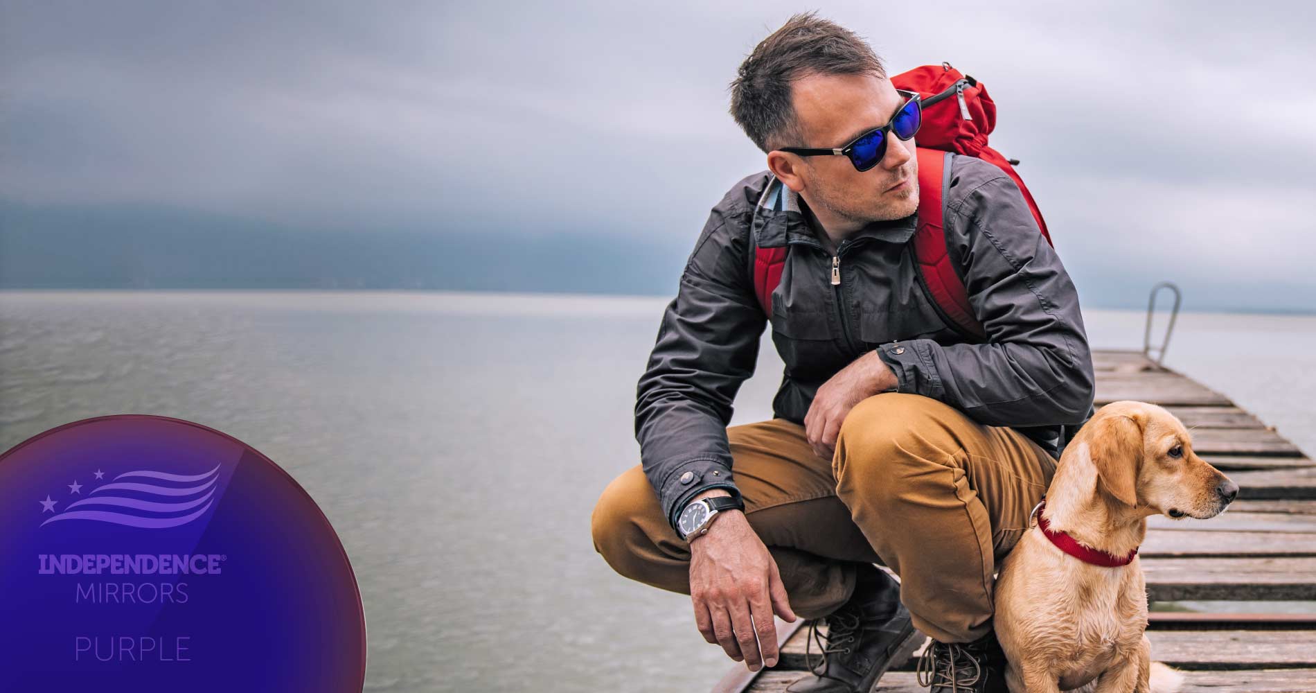 Man with hiking gear crouched down on a pier with a dog, wearing purple-colored mirrored sunglasses